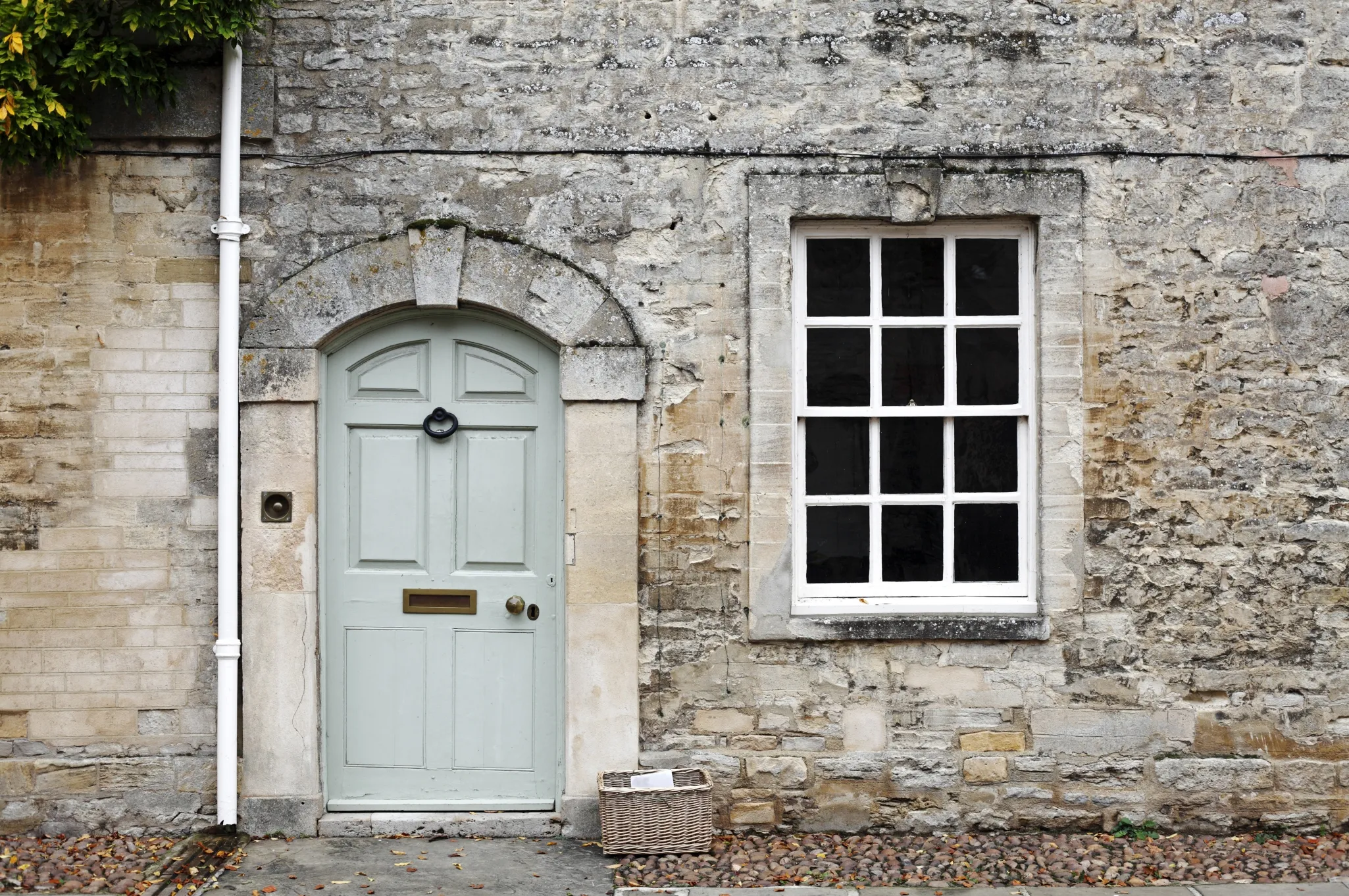 A stone house facade with a pale green door, an arched stone frame, a white-framed window, and a wicker basket on the ground.