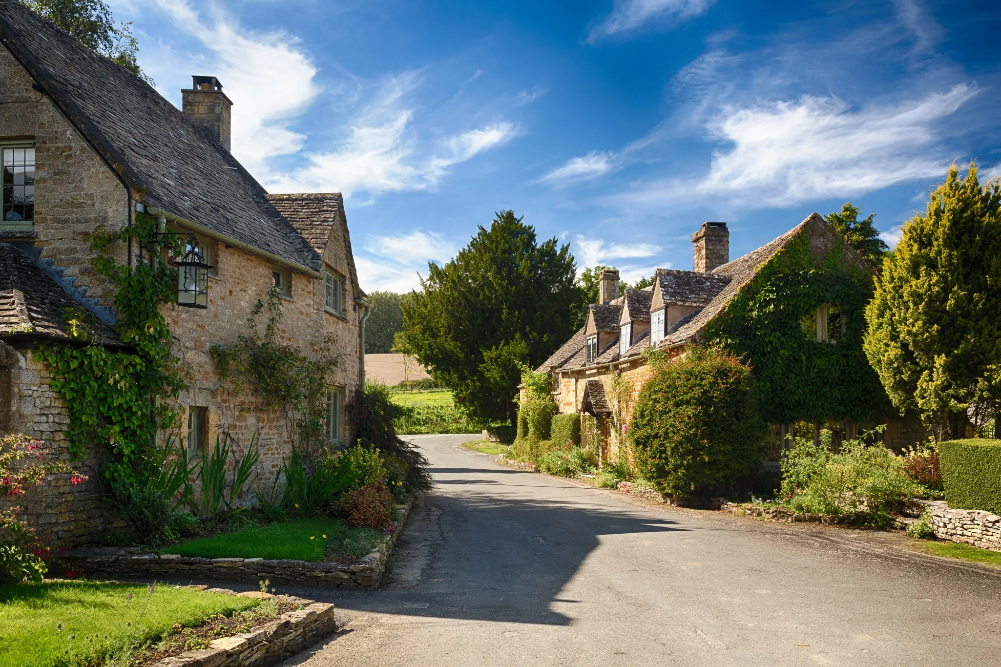 A narrow road with traditional stone cottages covered in greenery on a sunny day. Trees and bushes surround the buildings.