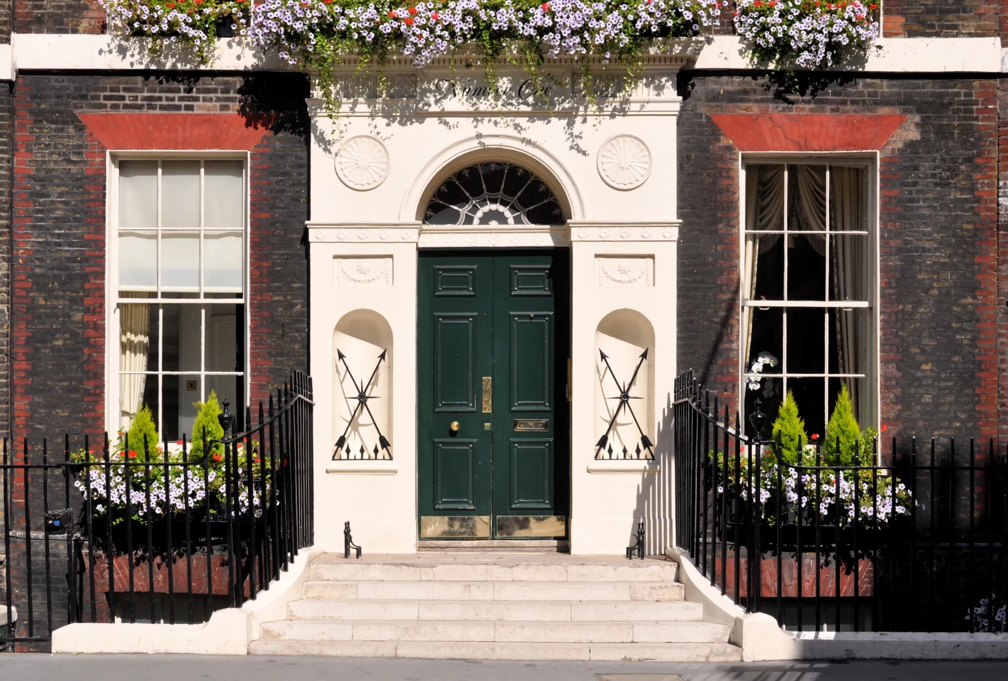 A black double door with a semicircular window above it, set between two large windows with flower boxes, flanked by brick walls.