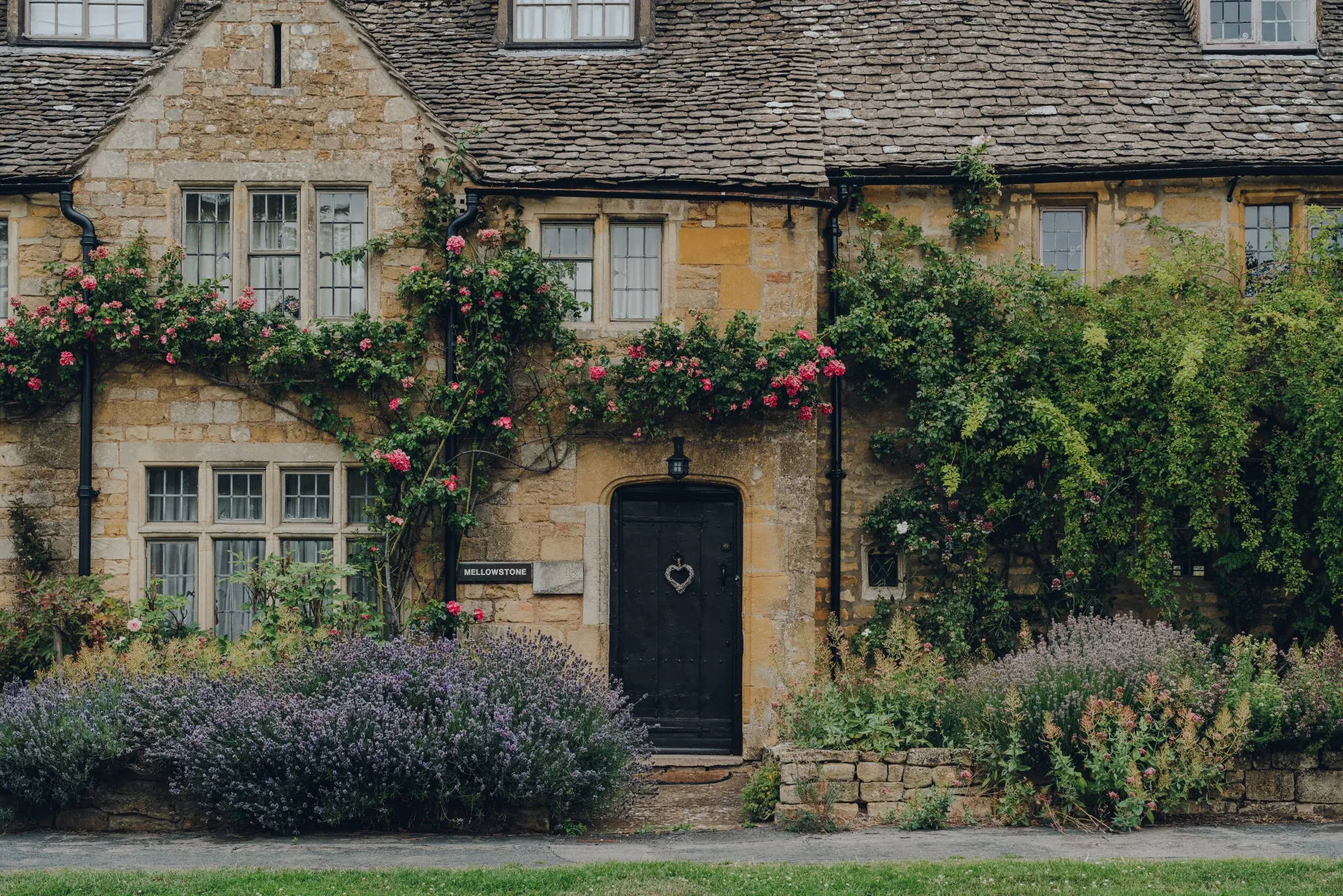 A stone cottage with a shingled roof, a black door with a heart decoration, and various flowering plants and vines around the façade.