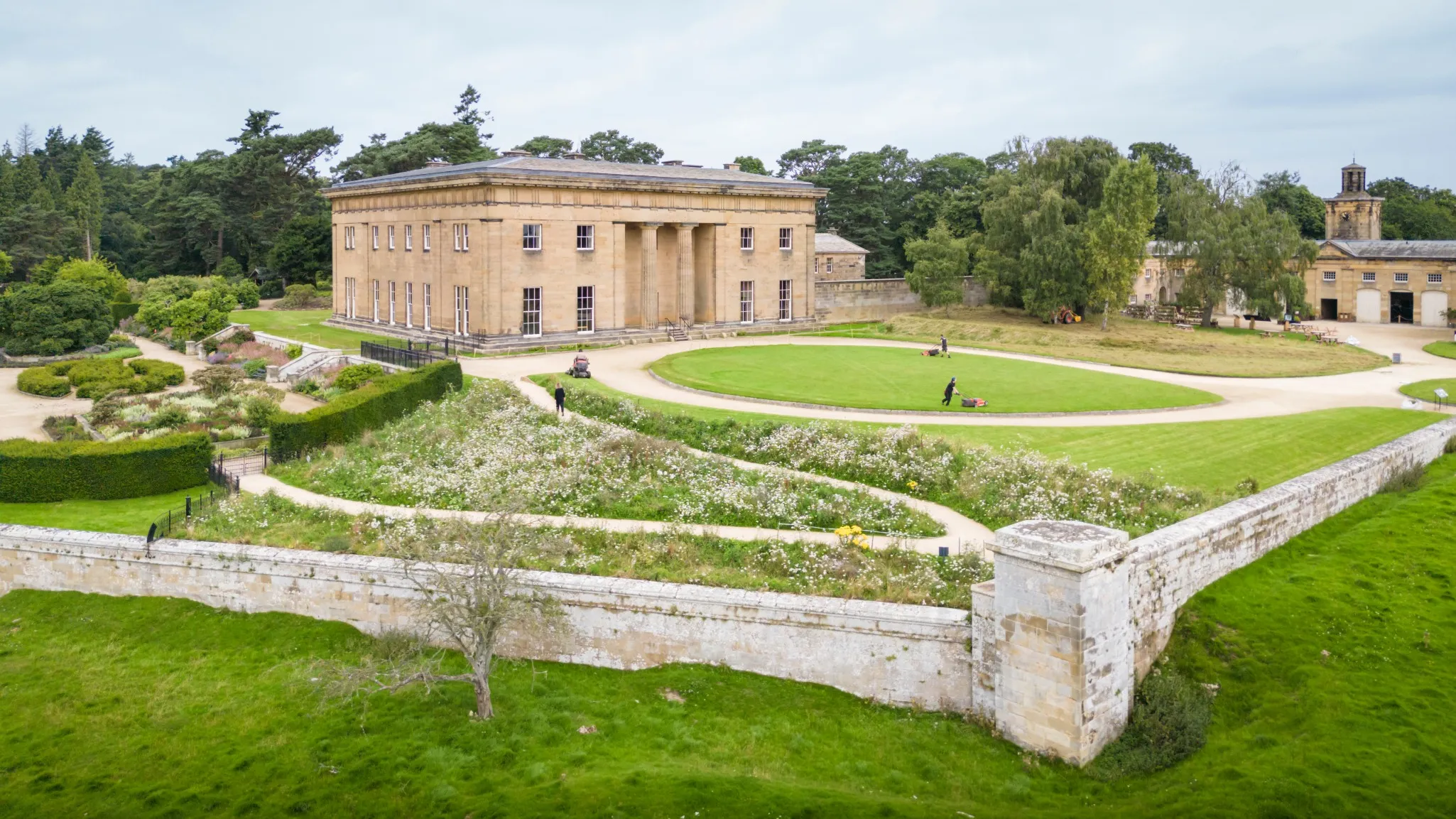 A large historic stone building with columns stands amidst expansive gardens and a circular driveway, surrounded by a low stone wall.