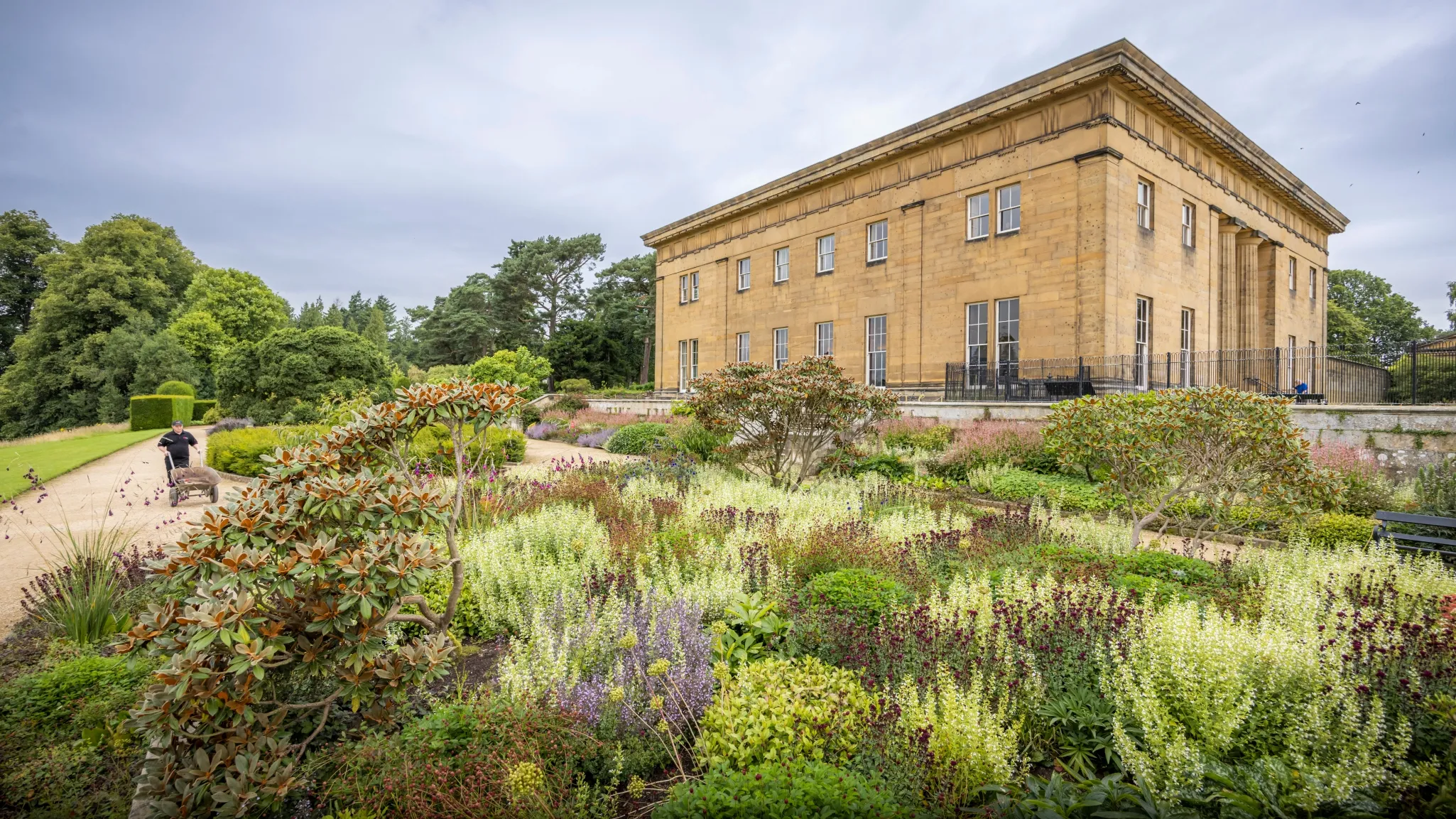 A large stone building with columns overlooks a garden filled with various plants and flowers on a cloudy day.