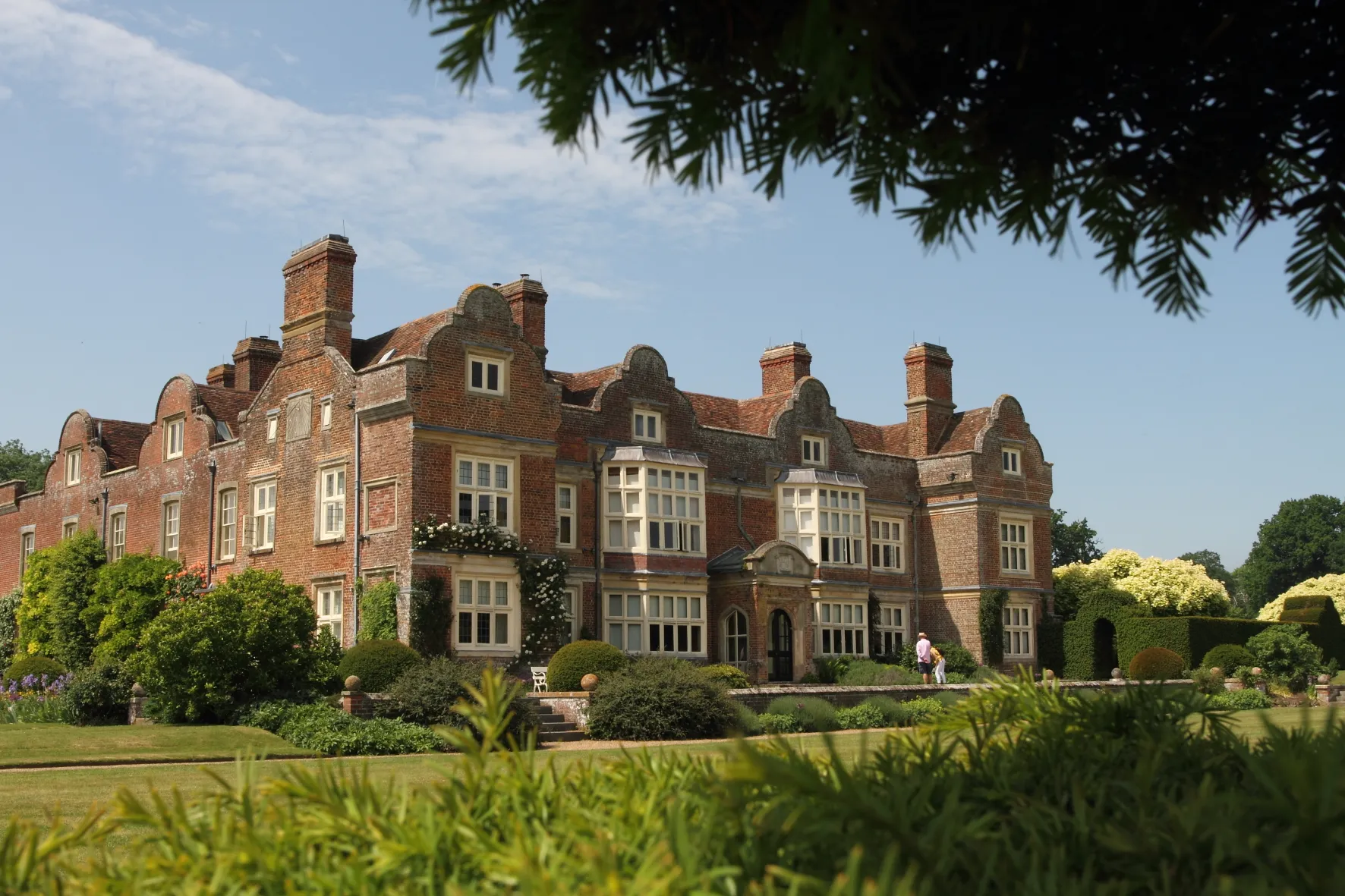 A large brick mansion with ornate windows and chimneys, surrounded by lush gardens and trees under a clear sky.