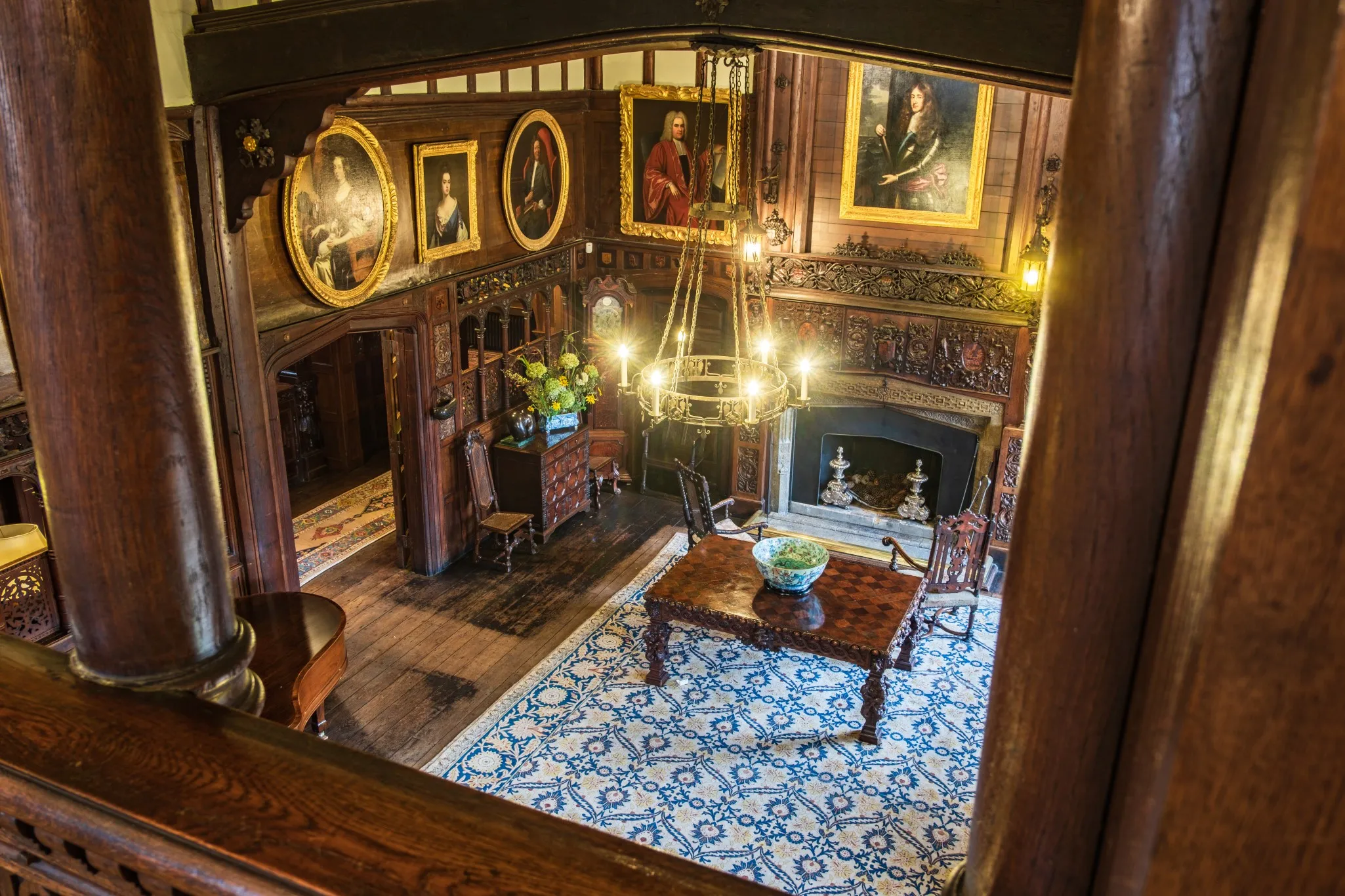 Ornate room with wooden walls, a large central table, paintings on the walls, a fireplace, chandelier, and patterned blue rug.