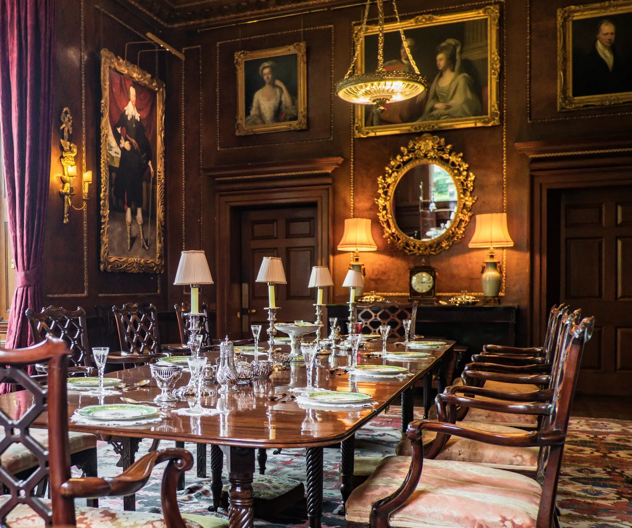 A classic dining room with an elegant wooden table set for a meal, surrounded by ornate chairs and decorated with portraits and lamps.