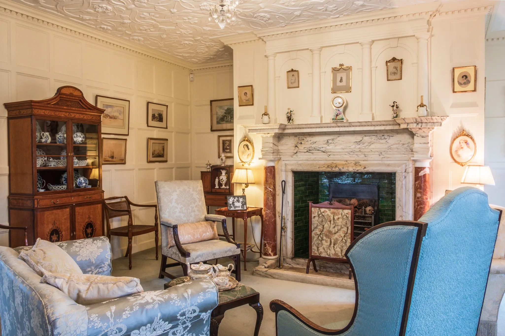 A vintage living room features ornate wall paneling, a decorative fireplace, antique furniture, framed art, and classic light fixtures.