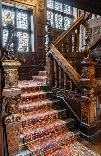 Intricately carved wooden staircase with ornate bannisters and patterned red carpet, leading up beside stained glass windows.