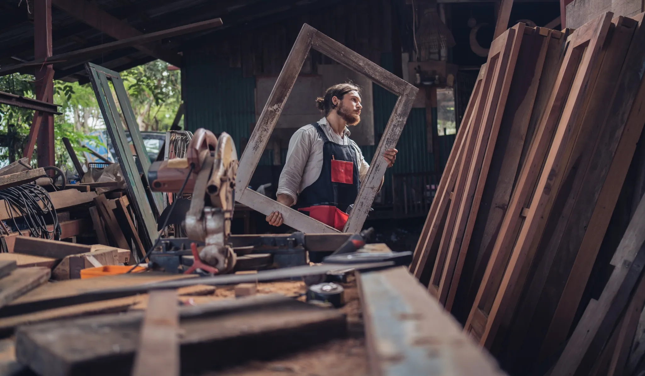 A person in an apron holds an empty wooden frame in a woodworking shop surrounded by wooden planks and tools.