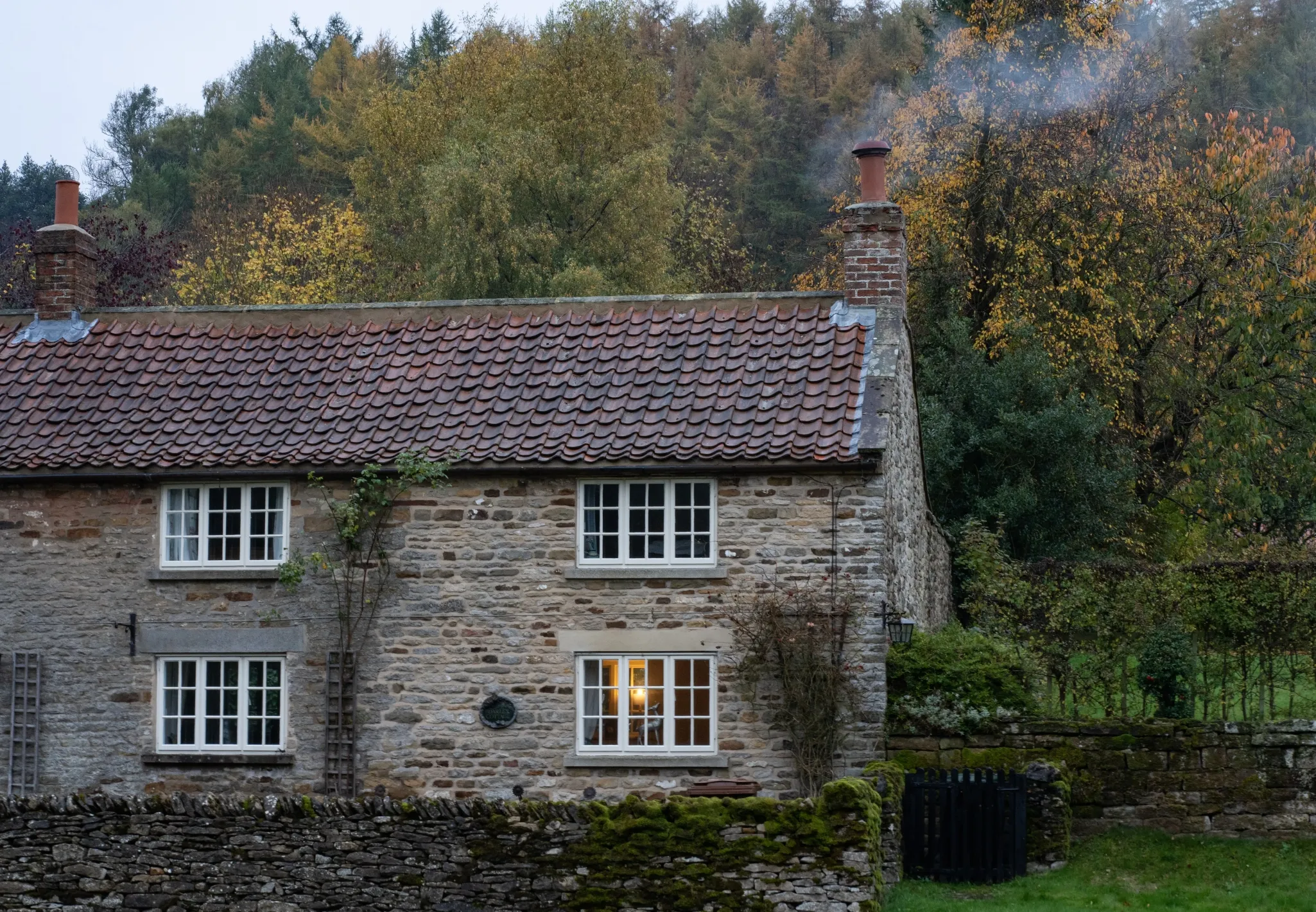 A quaint stone cottage with a tiled roof, surrounded by autumn foliage, has smoke rising from the chimney.