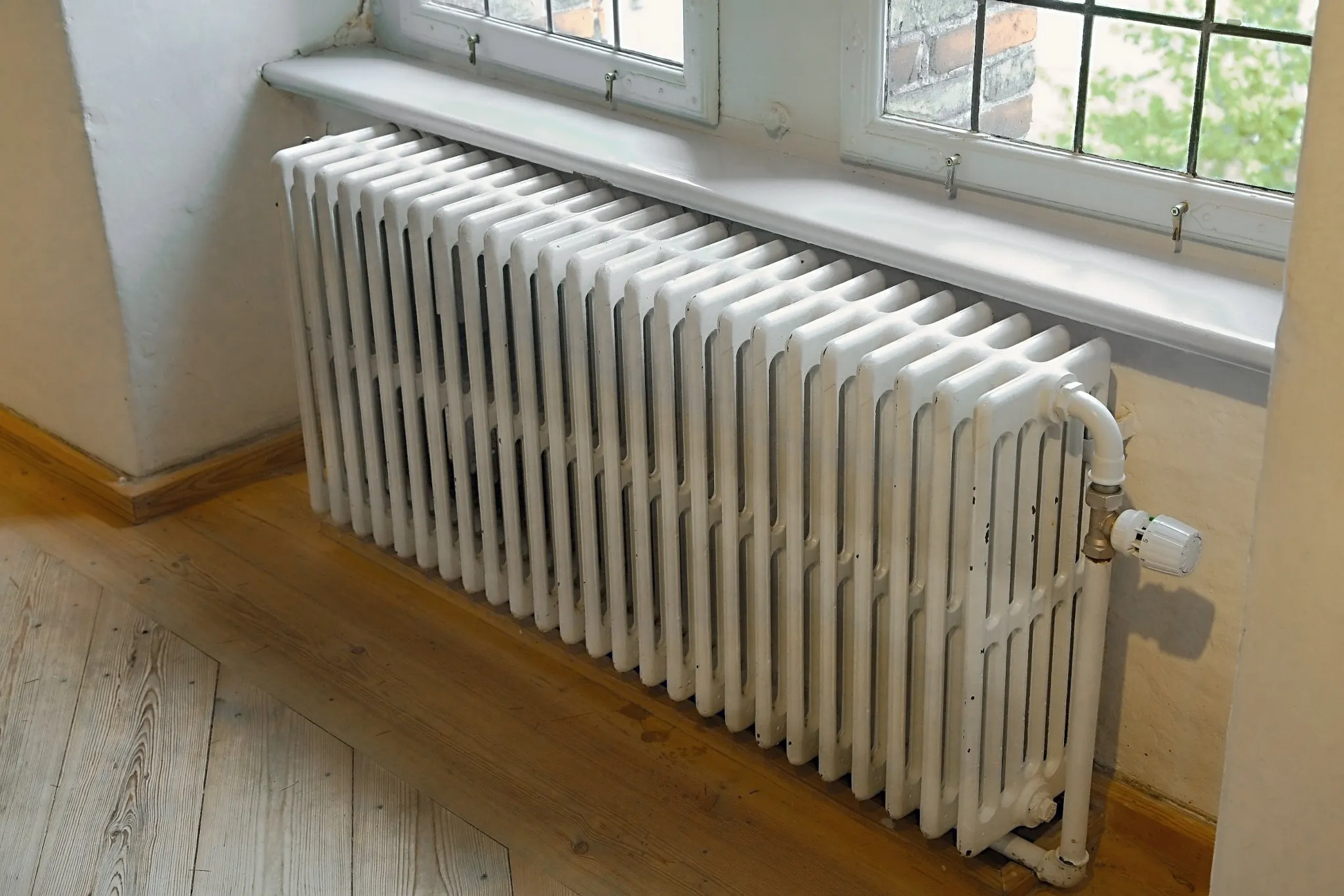 A white radiator placed beneath a pair of windows with wooden flooring surrounding it.