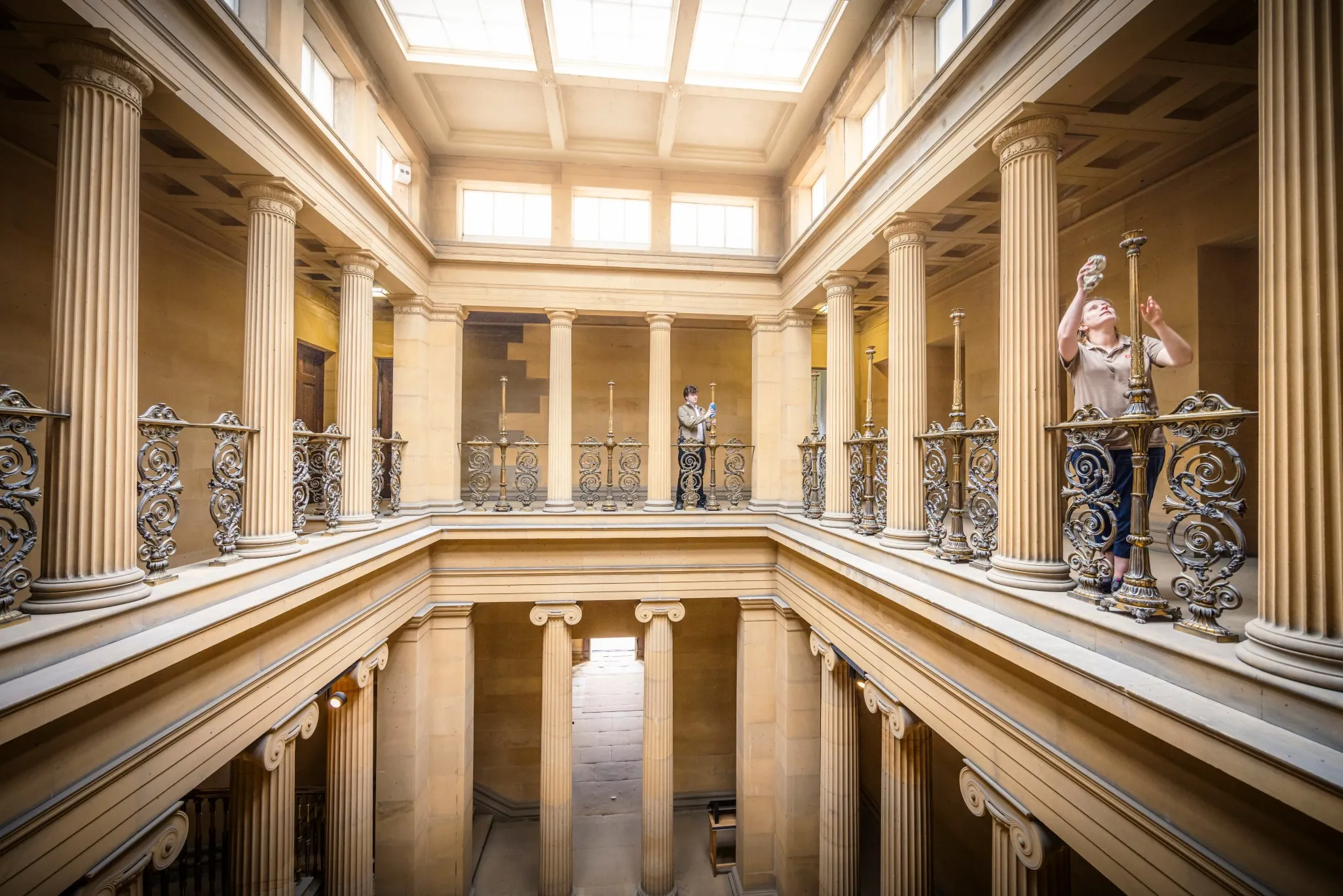 Two people cleaning stand on an ornate balcony with columns inside a large, well-lit building with a skylight.