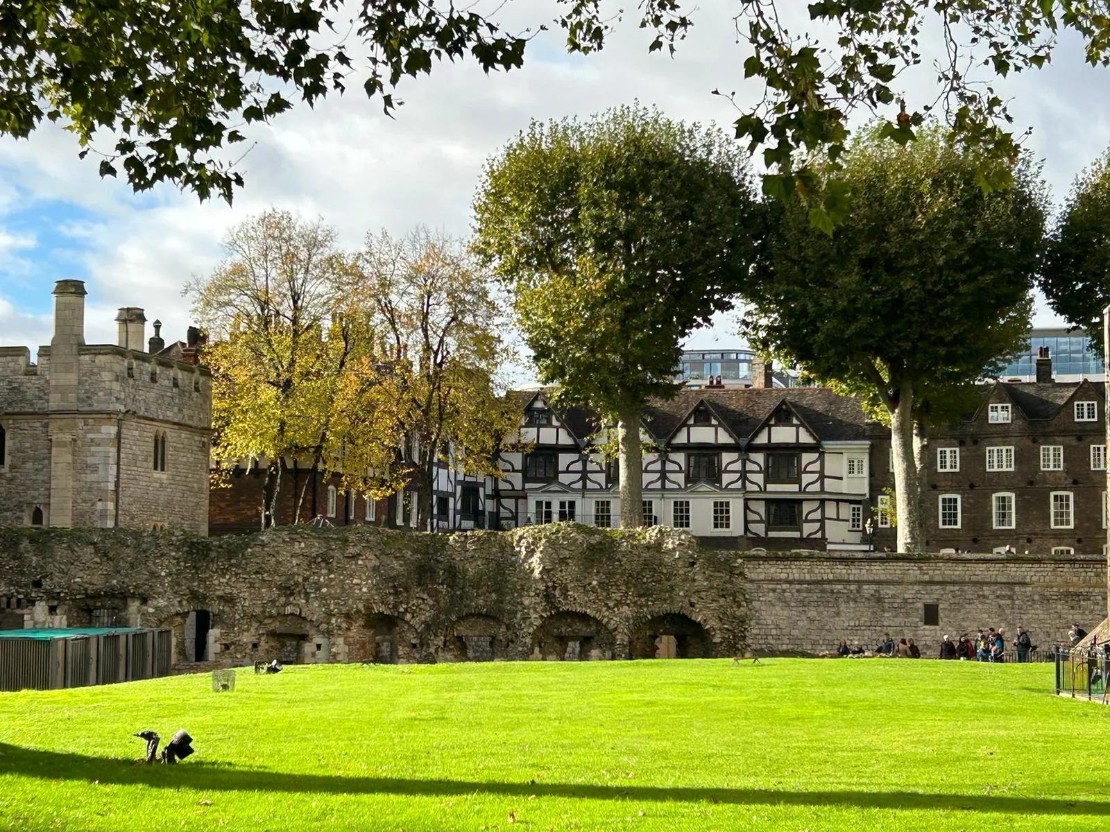 A historic stone building and timber-framed houses behind a large grassy area, with trees and scattered people around.