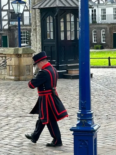 A man in a traditional uniform with red accents and a hat walks on a cobblestone path, passing a blue lamppost and old buildings.