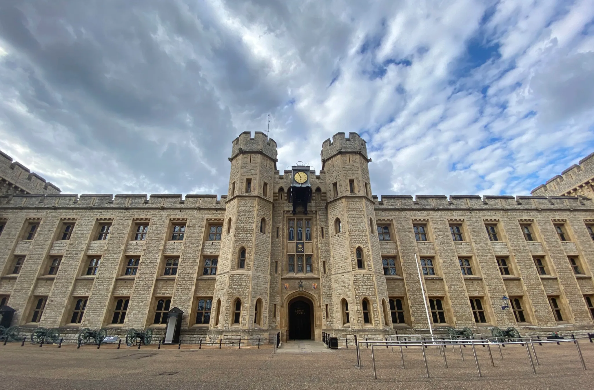 A historic stone castle with symmetrical towers and large clock sits under a cloudy sky, viewed from a courtyard.