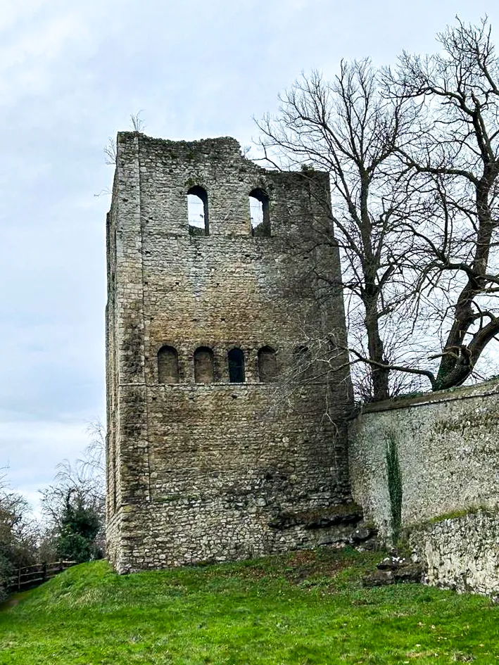 Stone tower of a historical building with arched windows, partially surrounded by a stone wall, bare trees in the background.