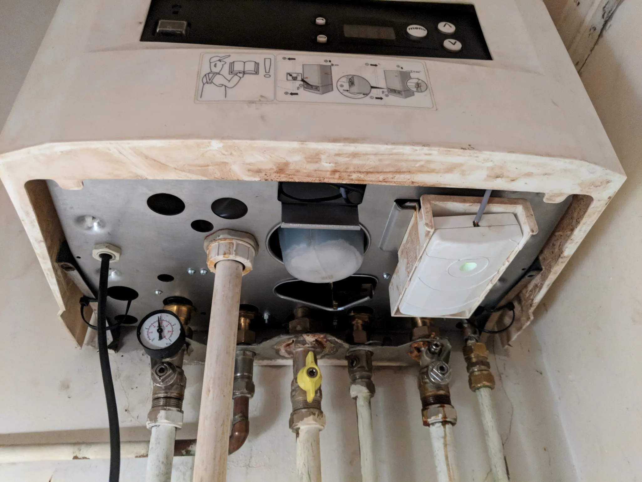 View of the underside of a boiler showing pipes, valves, a pressure gauge, and a thermostat, with visible signs of rust.