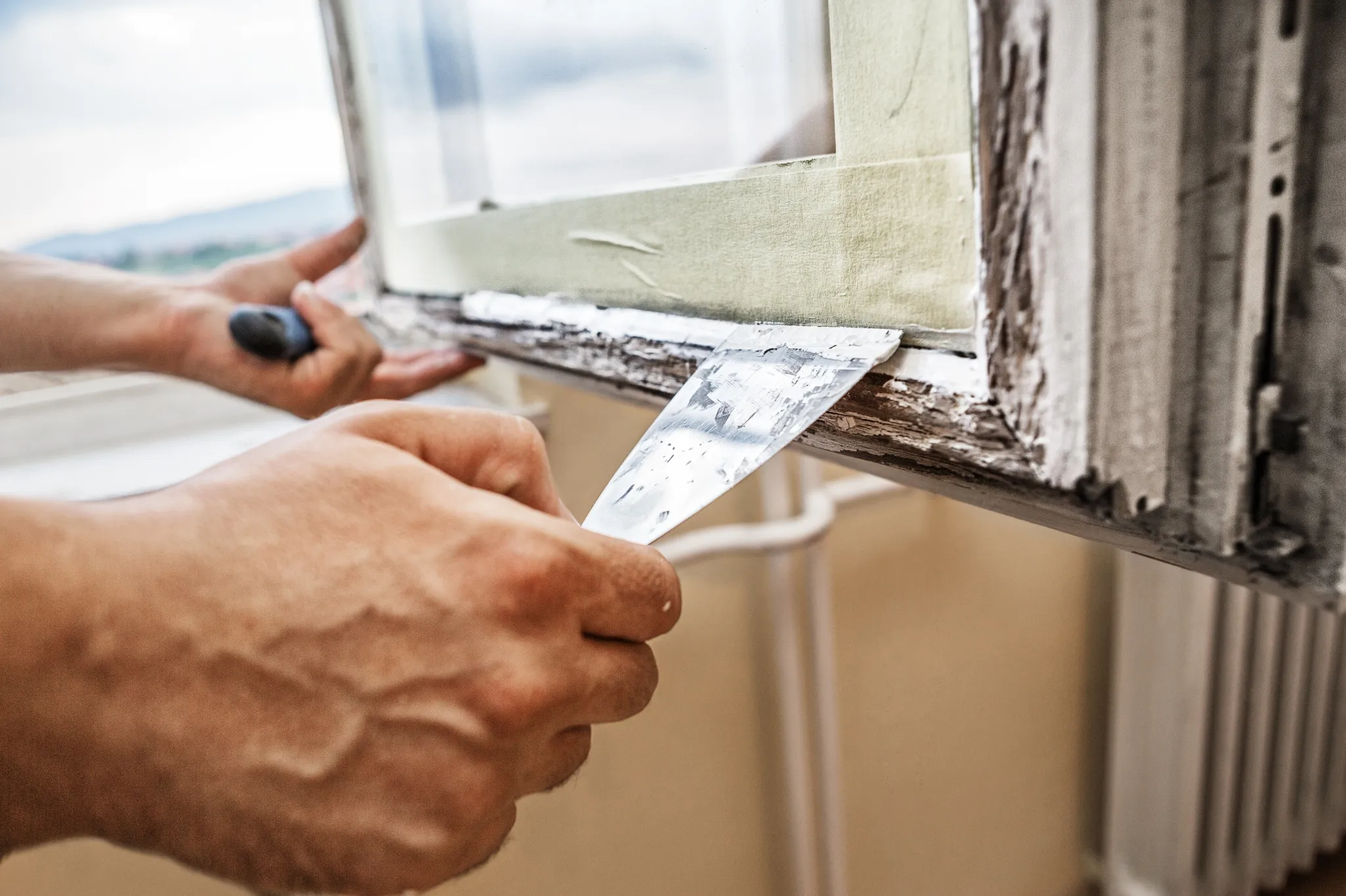 A person uses a putty knife to remove old paint from a window frame before repainting it.