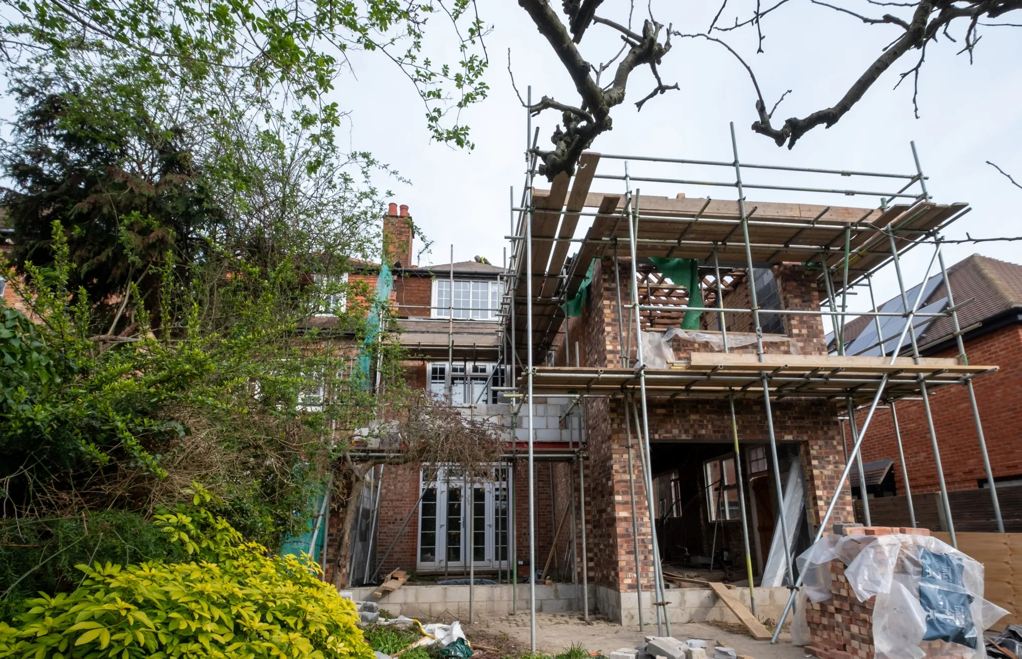 A two-story brick house under renovation, with scaffolding and construction materials visible around the site.