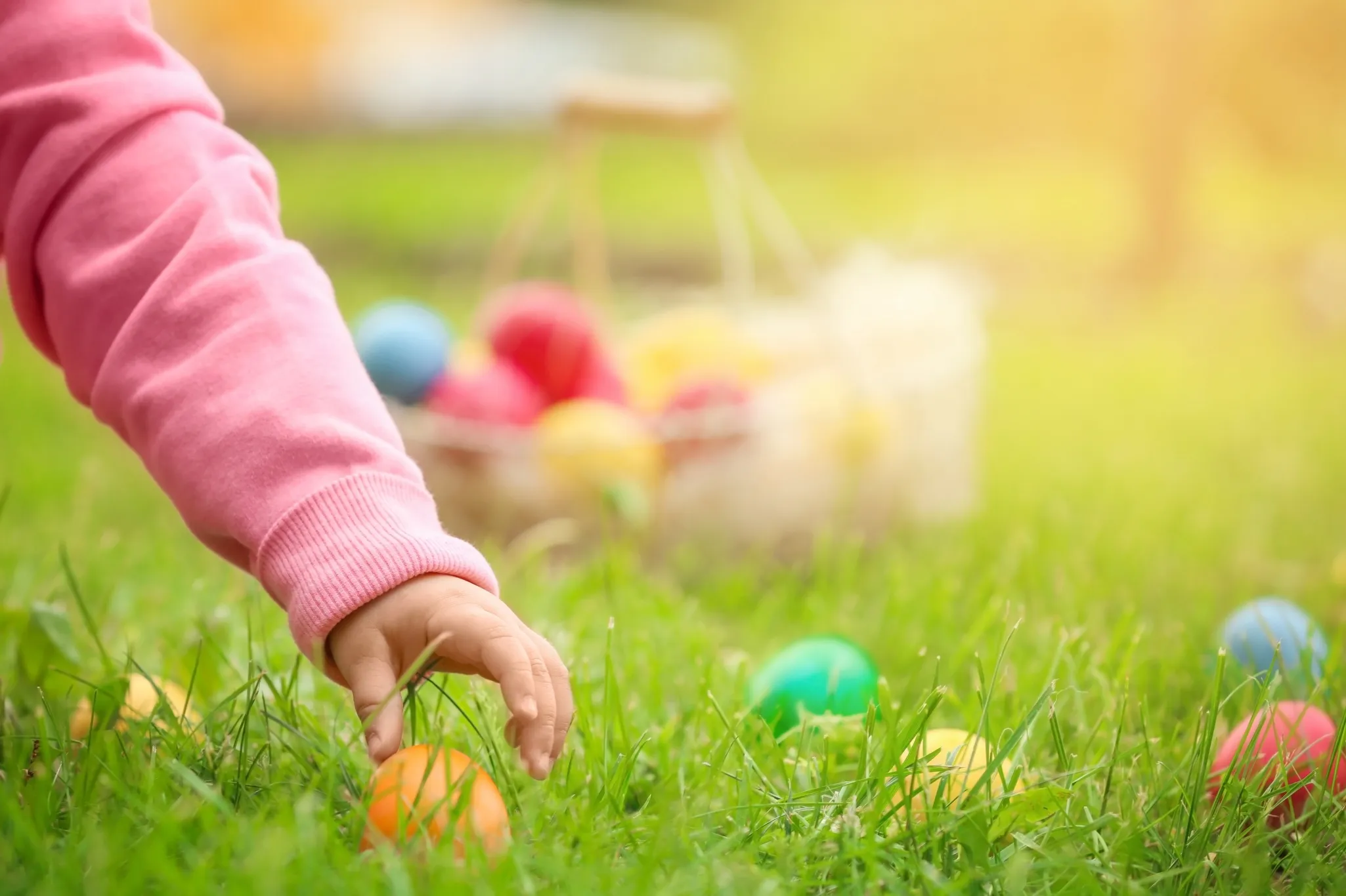 A child's hand reaching for a colorful egg on grass, with a basket of eggs in the blurred background.