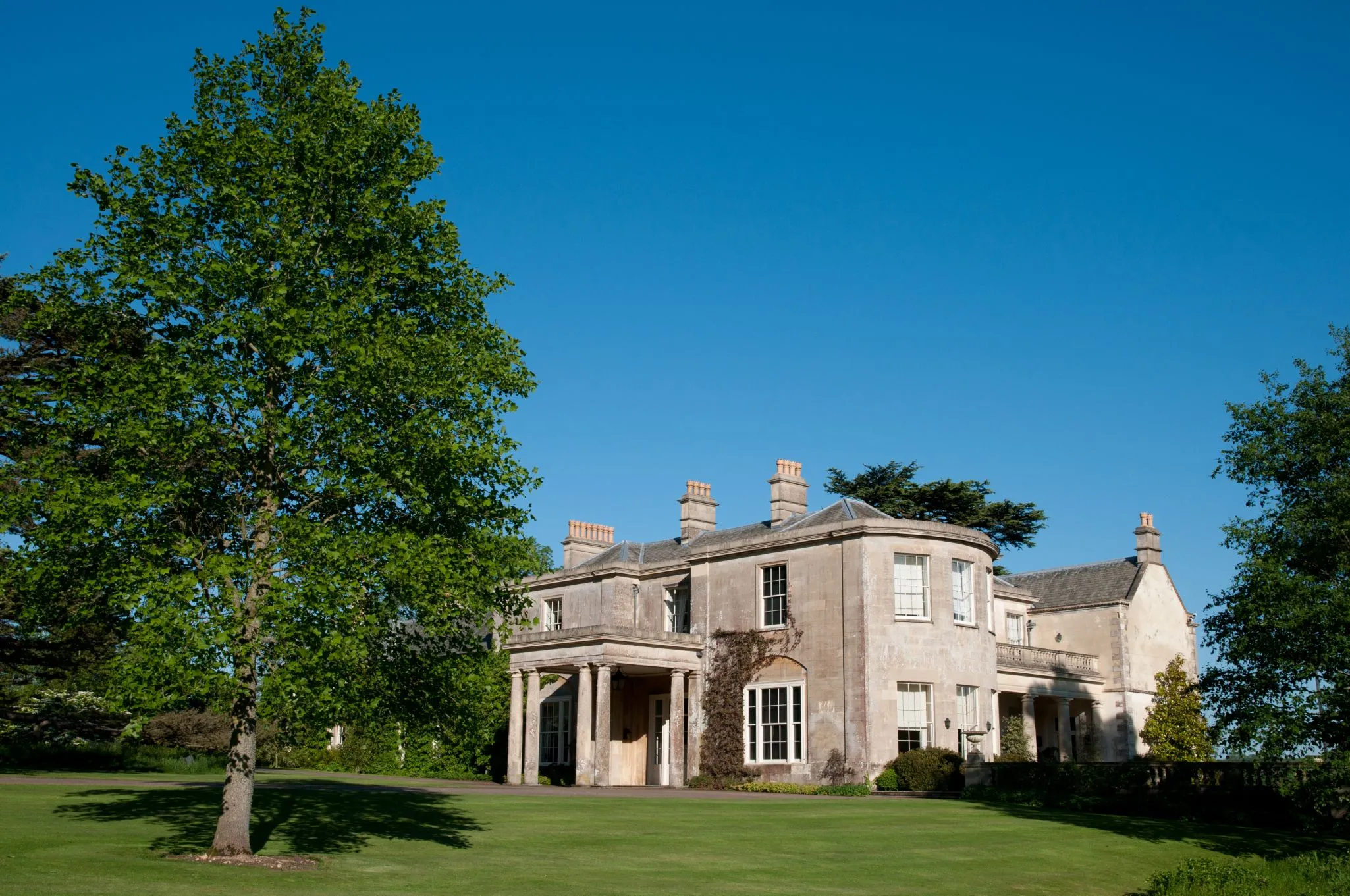 A large, historic mansion with multiple chimneys and columns, surrounded by lush greenery and trees, under a clear blue sky.