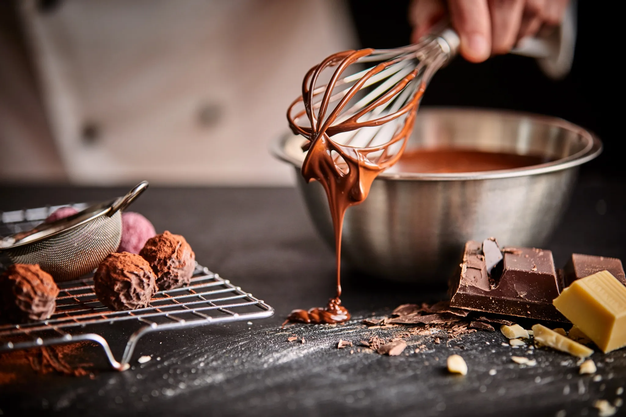 A person uses a whisk to mix melted chocolate in a bowl. Nearby are truffles, a strainer, and chunks of dark and white chocolate.