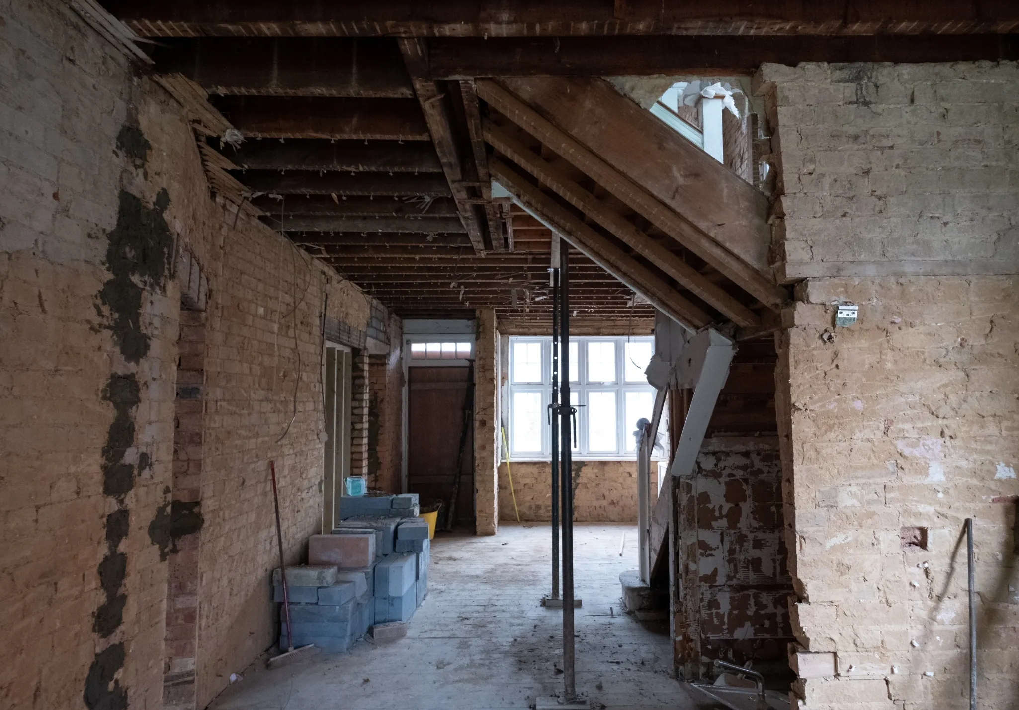 Partially demolished interior of a house with exposed brick walls, ceiling beams, and construction materials scattered.