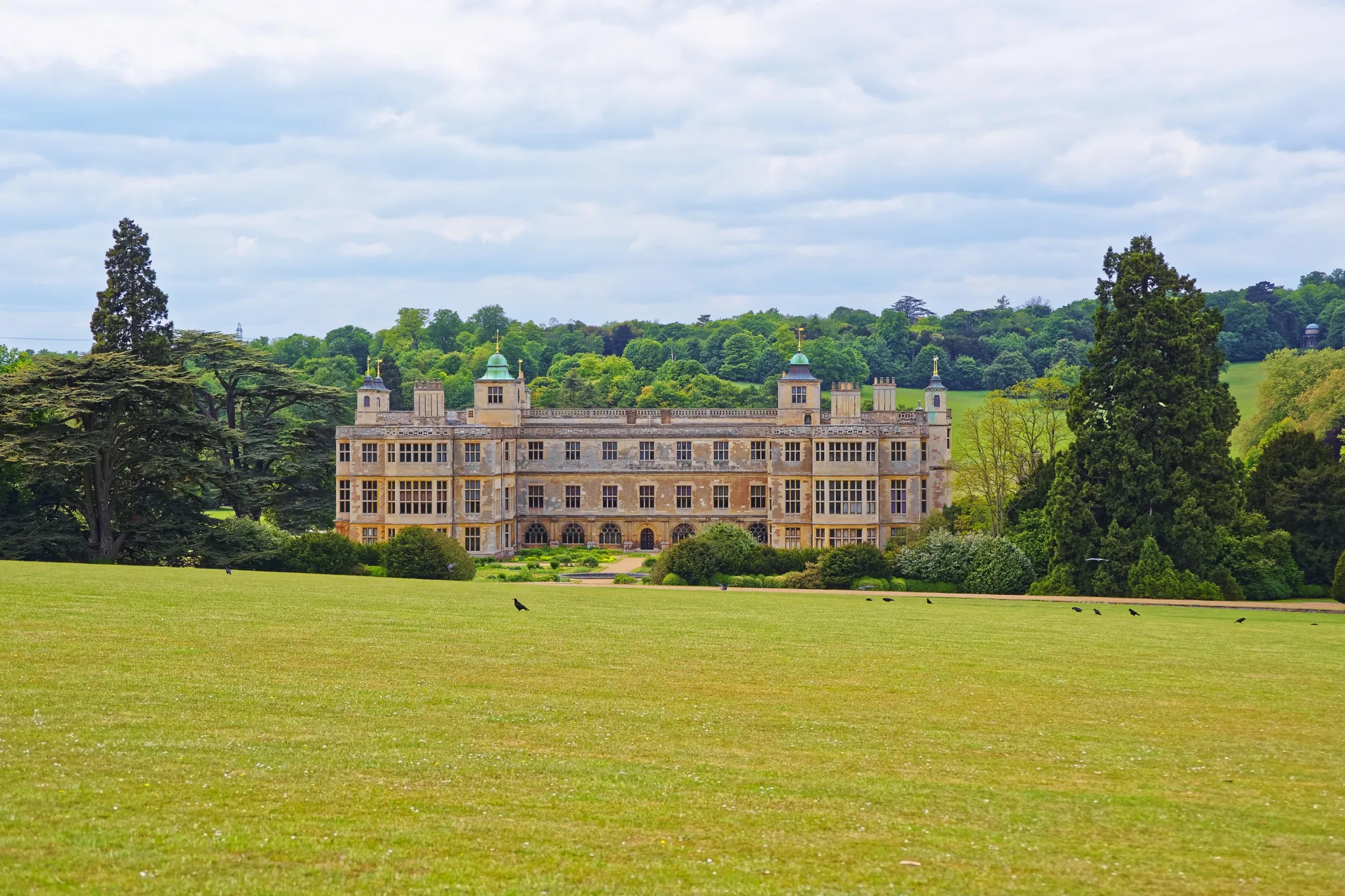 Large historic mansion with multiple windows and towers, set amidst expansive green lawns with trees and a partly cloudy sky.