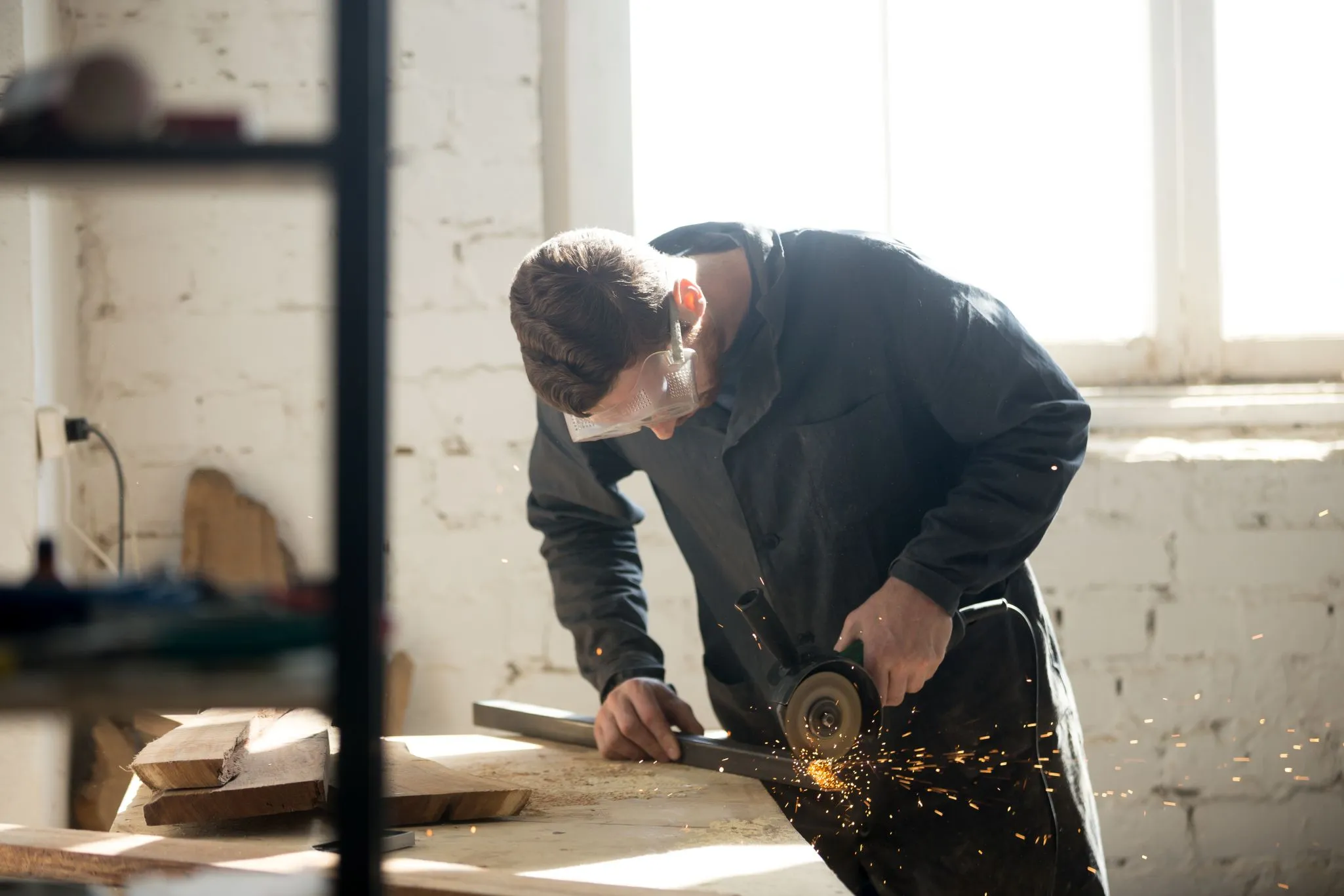 A person wearing safety goggles uses a grinder on a metal bar in a workshop, with sparks flying from the tool.