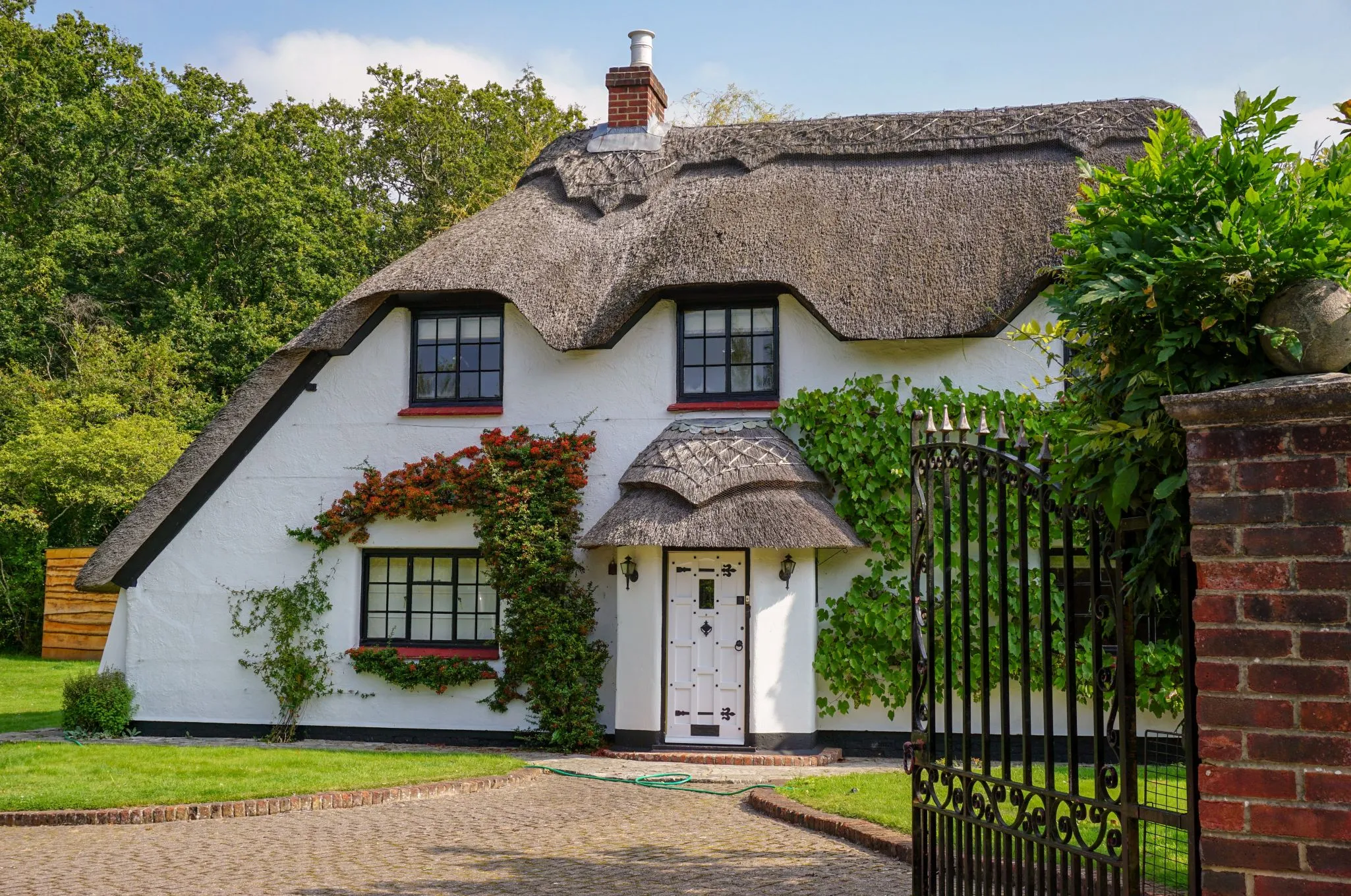 A quaint white cottage with a thatched roof, surrounded by greenery, and a wrought-iron gate in the foreground.