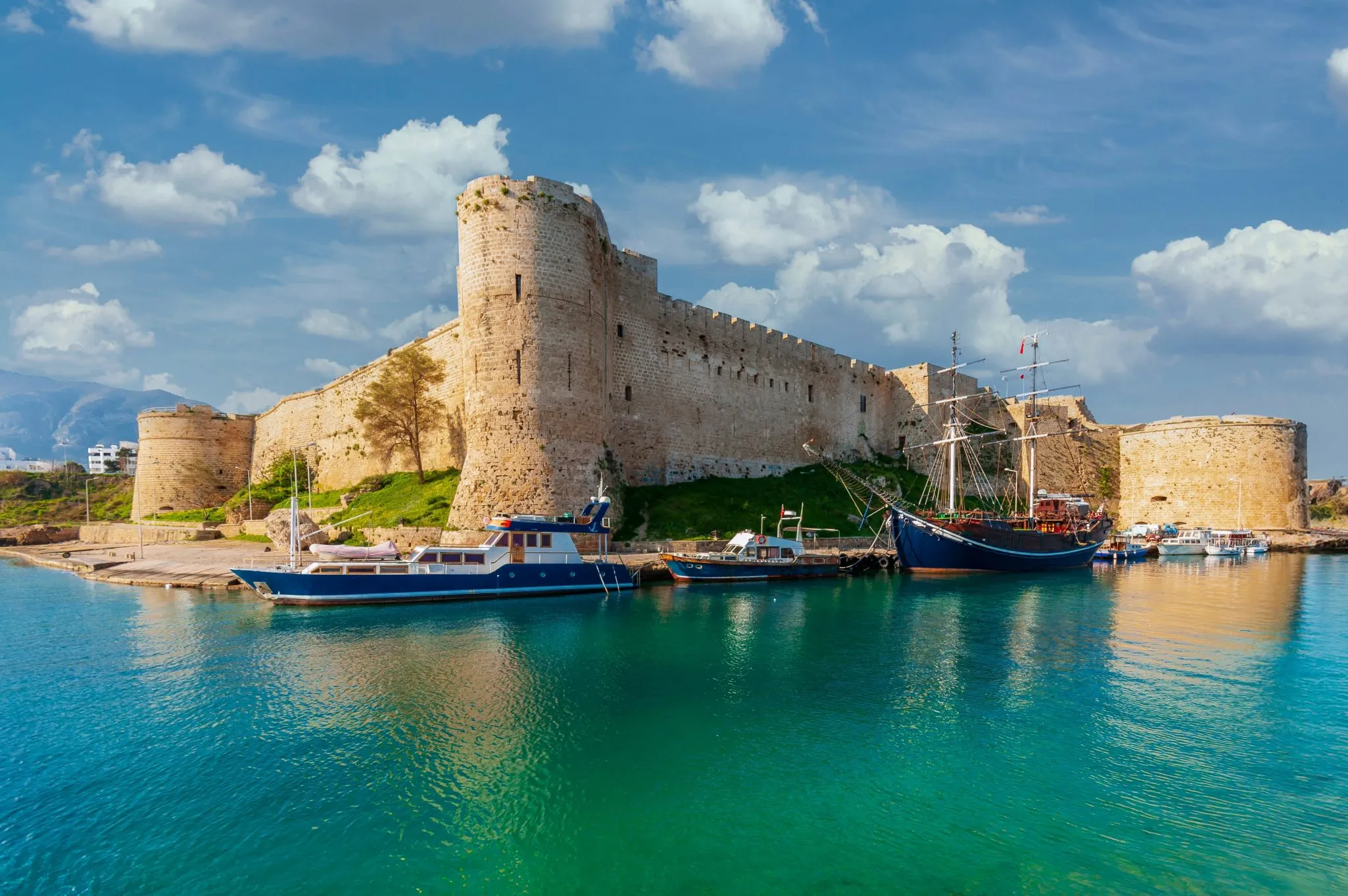 Medieval fortress with stone walls next to a calm, blue harbor with several docked boats under a partly cloudy sky.