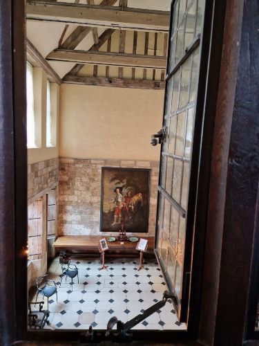 View through a window of a historic room with wooden beams, a large painting, a checkered floor, and a few pieces of furniture.