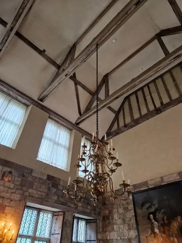 A grand, vintage chandelier hangs from a high ceiling with wooden beams in a room featuring stone walls and large windows.