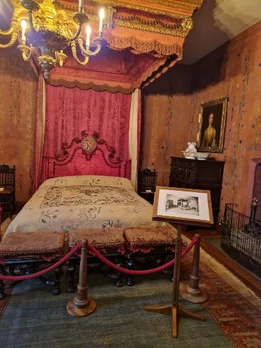 A furnished historical bedroom features an ornate canopy bed, a chandelier, a portrait on the wall, and a stand with a framed photo.