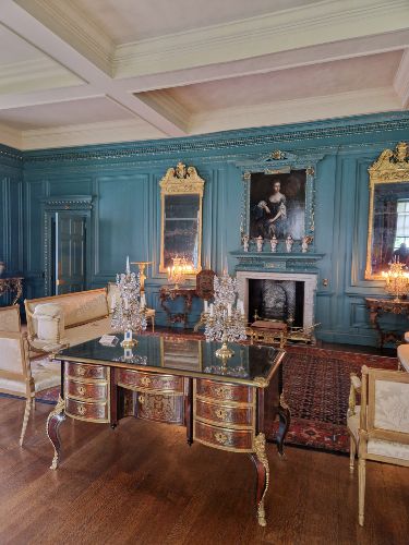 An opulent room with teal walls, ornate furniture, a desk in the center, large mirrors, a fireplace, and a portrait above the mantel.