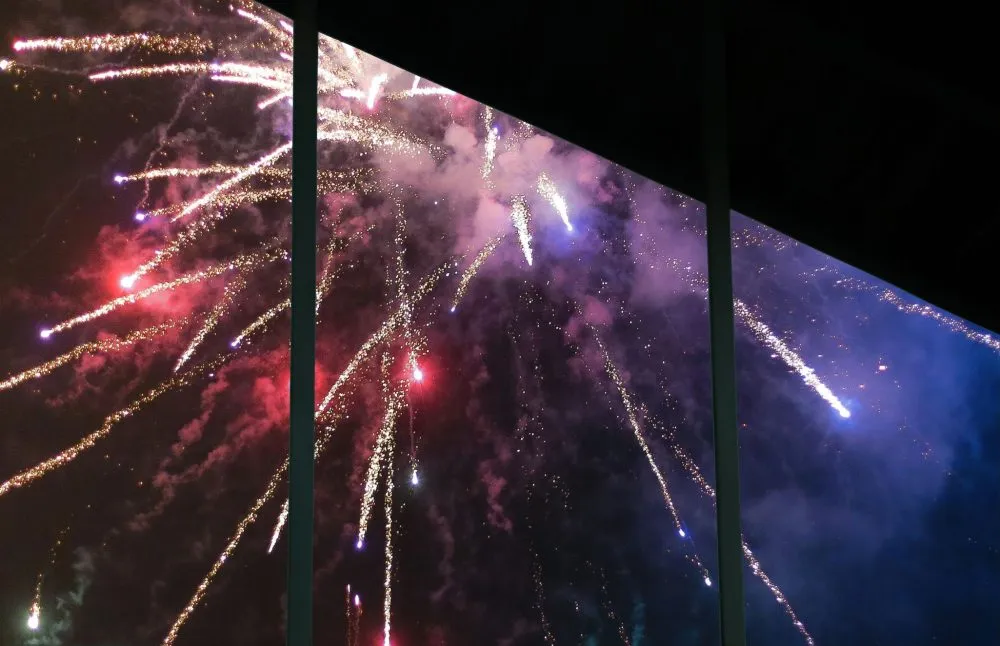 Colorful fireworks burst in the night sky, viewed through a window with vertical black bars.