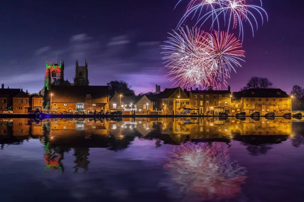 Fireworks light up the night sky over a town with illuminated buildings reflecting on the calm water.