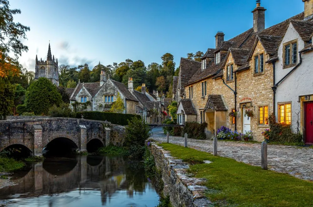 A picturesque village scene with stone cottages, a stone bridge, and a small river under a twilight sky.