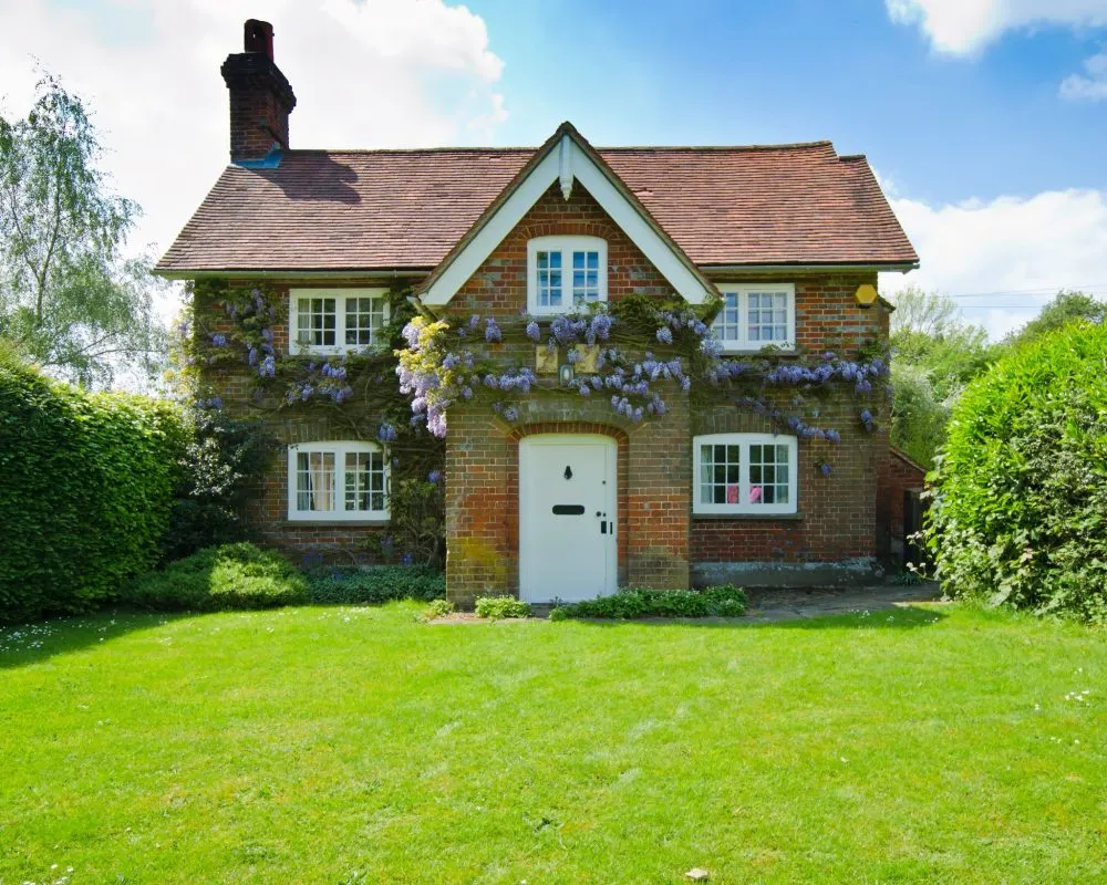 Charming brick cottage with lush vines and a red roof, framed by greenery and under a blue sky.