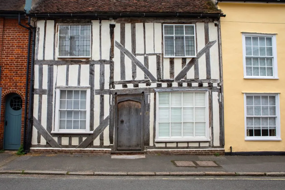 A historic timber-framed house with a wooden door and white windows stands between brick and plaster buildings on a street.