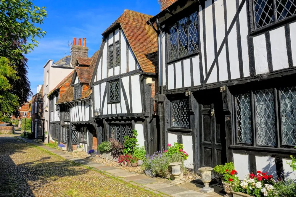 A row of traditional black and white timber-framed houses with flower pots and a cobblestone street under a clear blue sky.