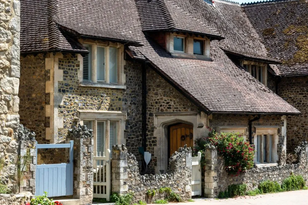 Stone house with a sloped roof, wooden door, windows with shutters, and a stone wall with foliage in front.