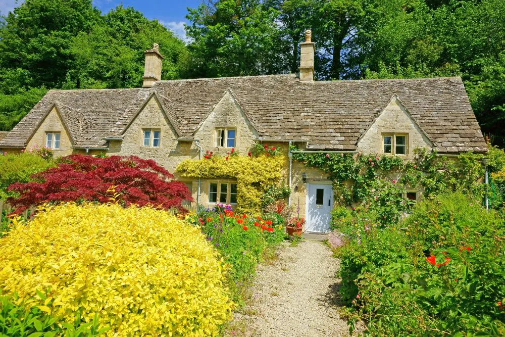 A stone cottage with a slate roof, surrounded by a colorful garden with various flowers and shrubs.