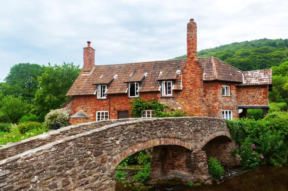 A rustic brick cottage with tiled roof and chimneys, behind a stone bridge, set in a lush green landscape under a cloudy sky.