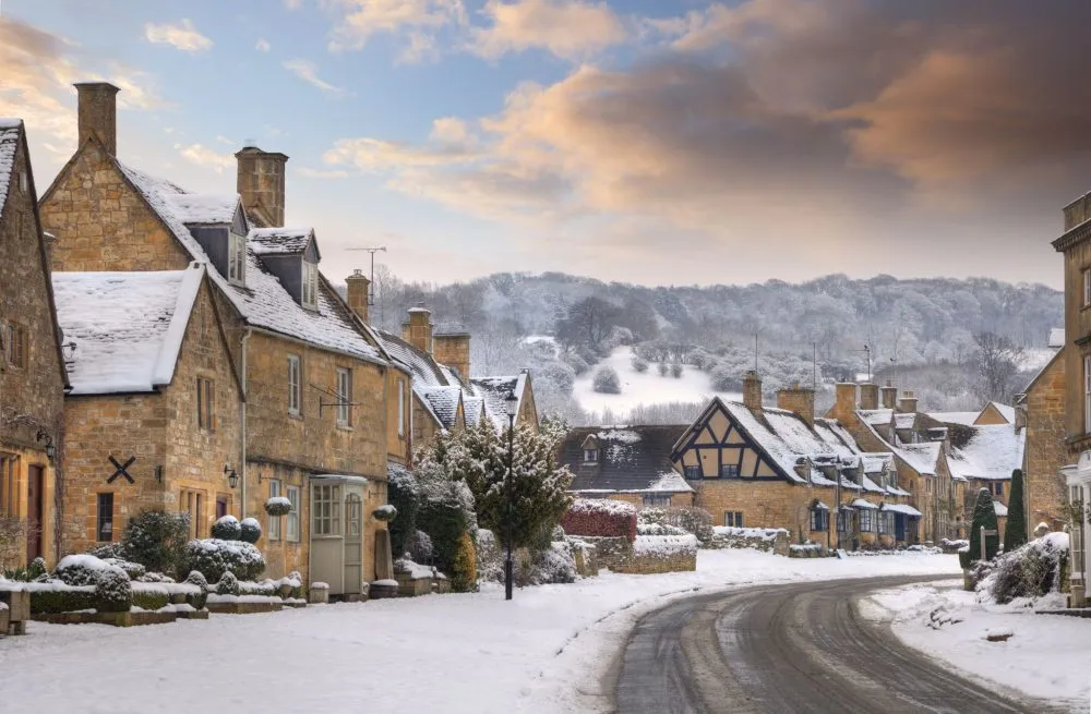 Quaint village street in winter with snow-covered stone houses and a backdrop of snowy hills under a partly cloudy sky.