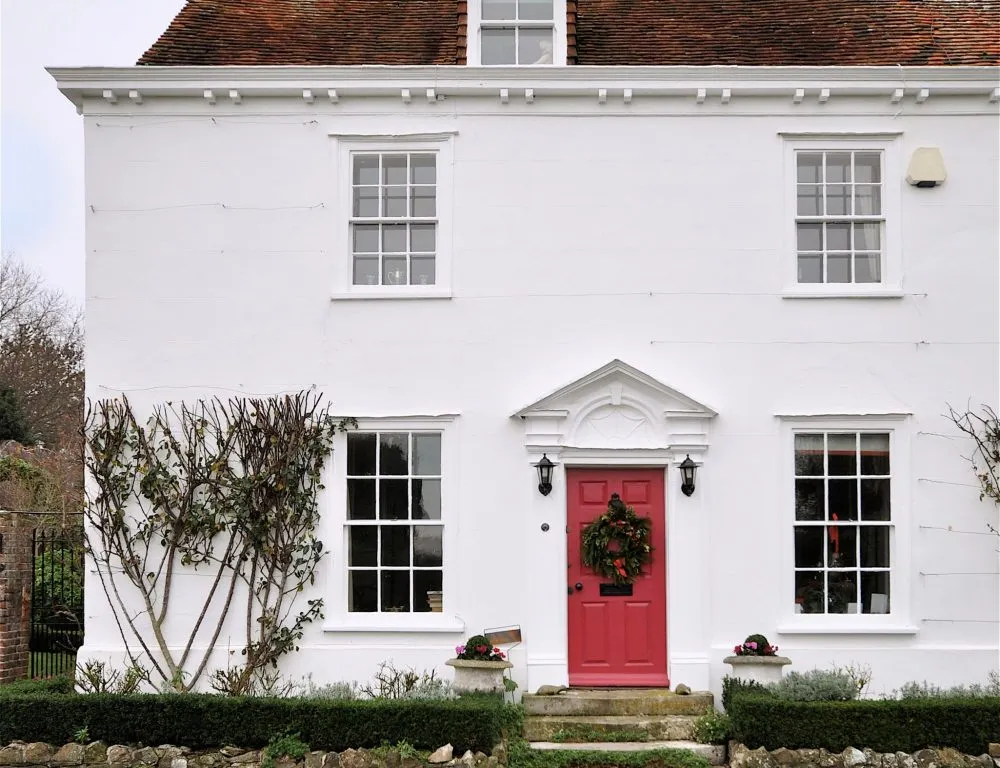 A white two-story house with a red door, decorated with a wreath, and trimmed bushes in the front.