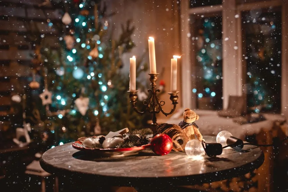 A candle holder and holiday decorations on a table, with a Christmas tree and softly falling snow in the background.