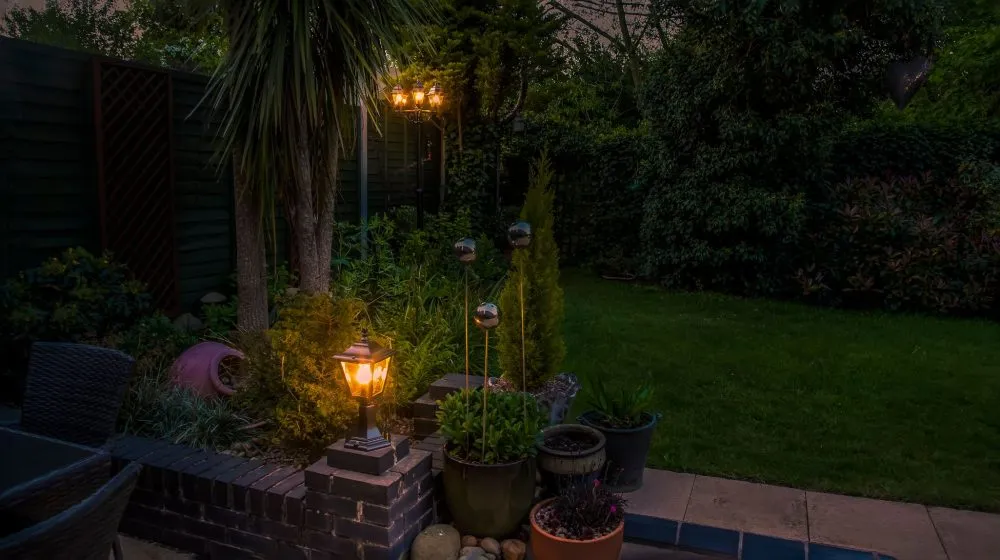 A garden at dusk with illuminated lanterns, potted plants, palm-like tree, and lawn surrounded by dense green foliage.