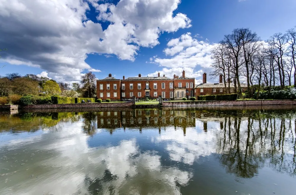 A large brick mansion with multiple chimneys, reflected in a calm pond, under a partly cloudy sky with surrounding trees.