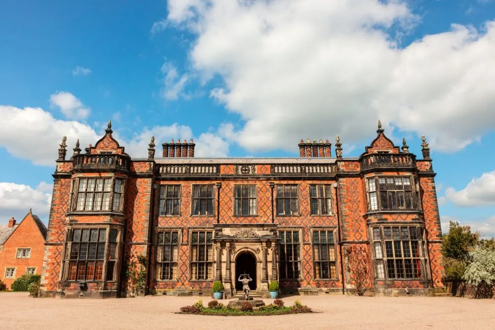 Historic brick mansion with intricate black detailing, central entrance, and large windows under a blue sky with clouds.