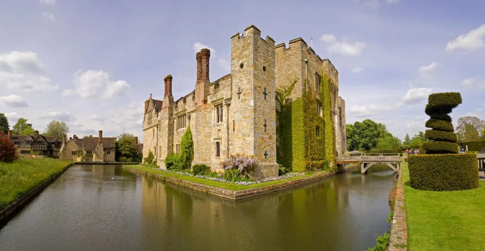 A historic stone castle with a moat and topiary gardens under a blue sky with clouds.